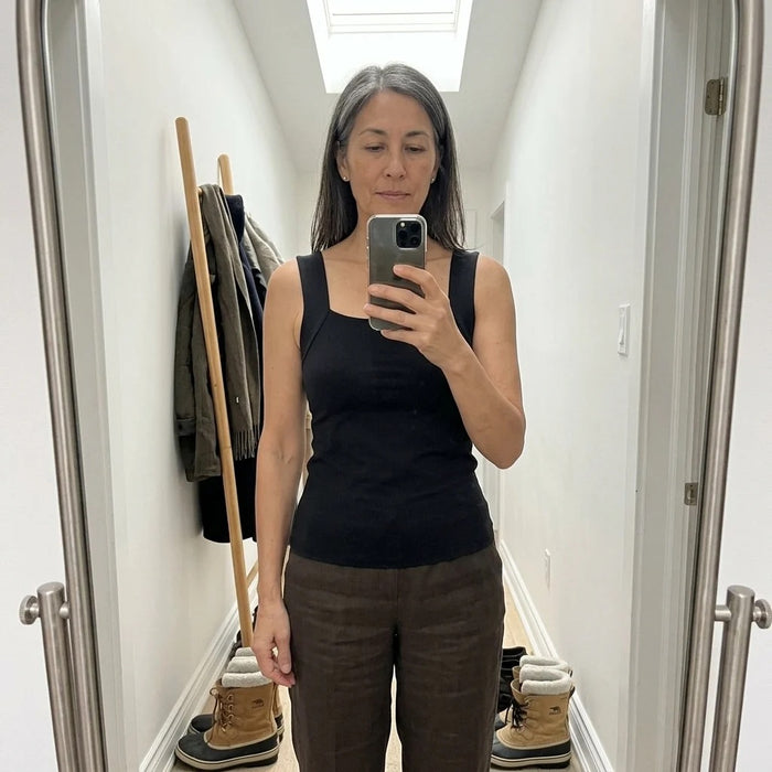 A woman in a bright hallway, wearing the Faye Built-In Support Tank in Black with linen trousers