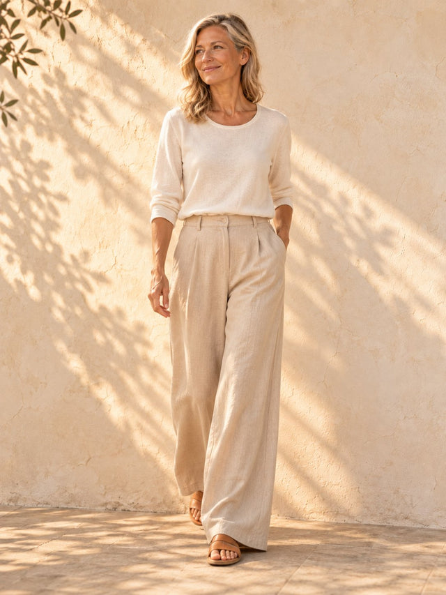 A woman in wide sand-coloured trousers and a simple top standing beside a sunlit plaster wall